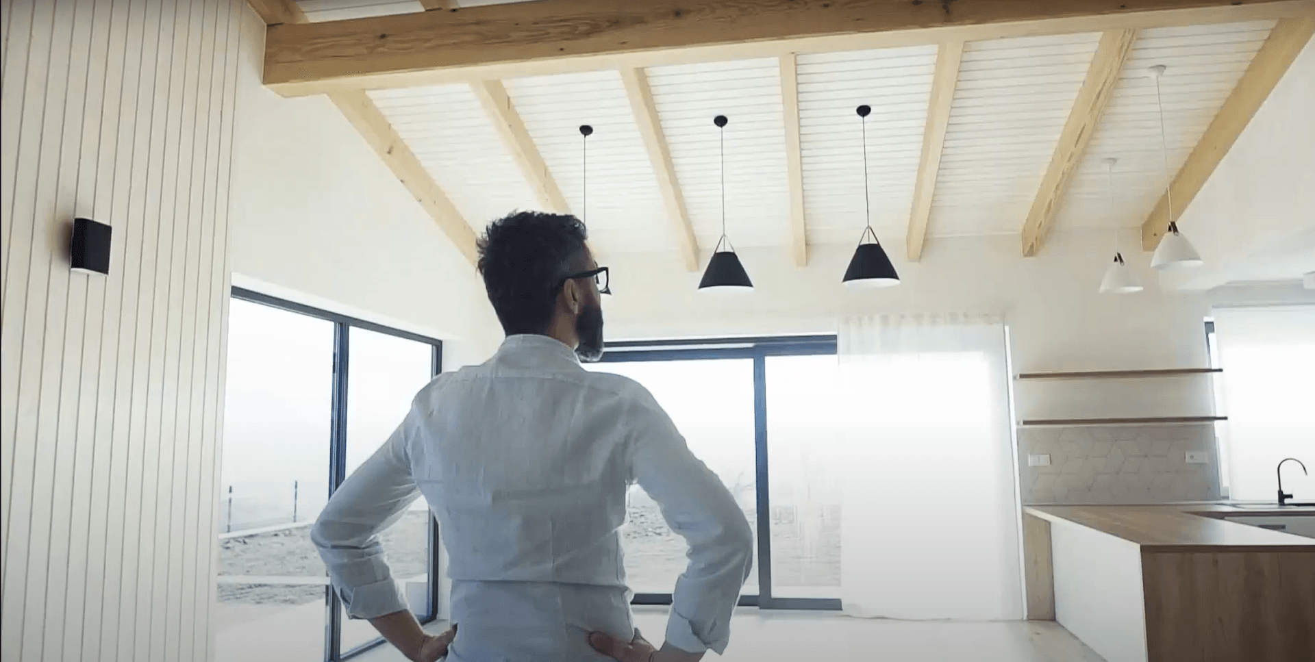 Person standing in a bright modern room with exposed beams and large windows