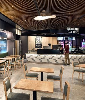 Modern café interior with wooden tables, chairs, curved sofa seating, and a person standing near the counter