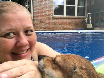 A person smiles while resting their chin on a pool float next to a wet brown dog in a backyard swimming pool.