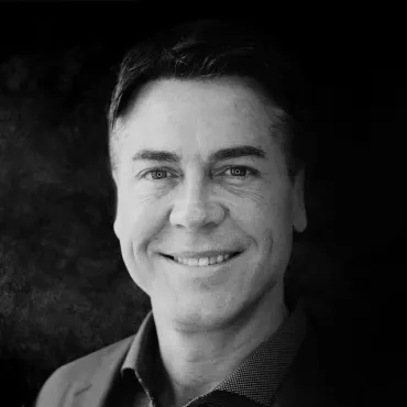 A headshot of a smiling person wearing a collared shirt against a dark, textured background.