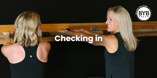 Two individuals in black athletic wear smile while exercising at a wooden ballet barre against a black background.