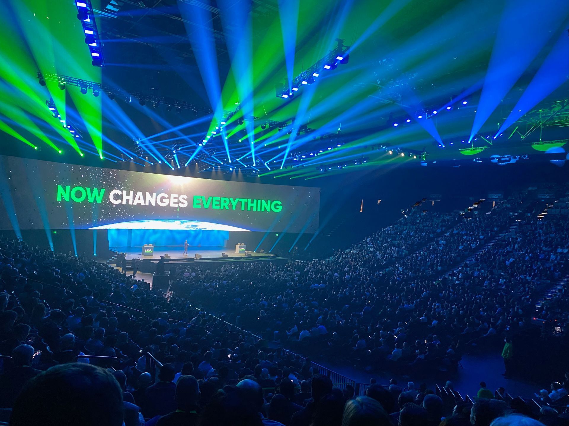 Concert arena with blue and green lasers and a stage screen reading “NOW CHANGES EVERYTHING”