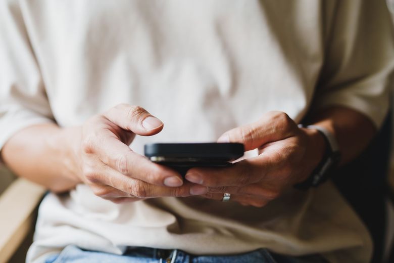 Person holding a smartphone with both hands while sitting indoors, wearing a beige shirt and jeans