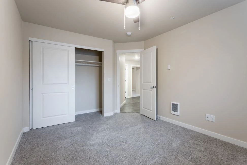 Empty carpeted bedroom with open closet and doorway to hall, beige walls, and ceiling fan light