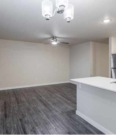 Empty modern living room with gray flooring, white walls, ceiling fan, and kitchen counter