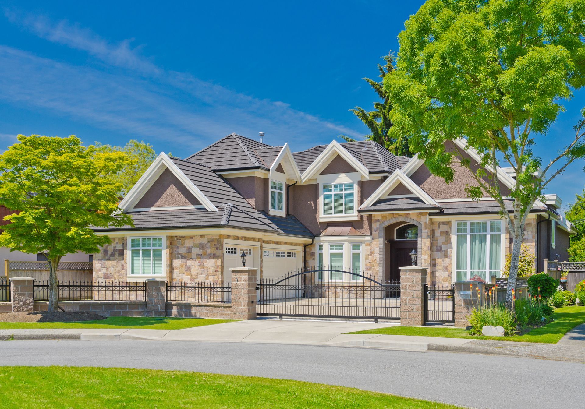 Large stone suburban house with a gated front yard, manicured lawn, and blue sky