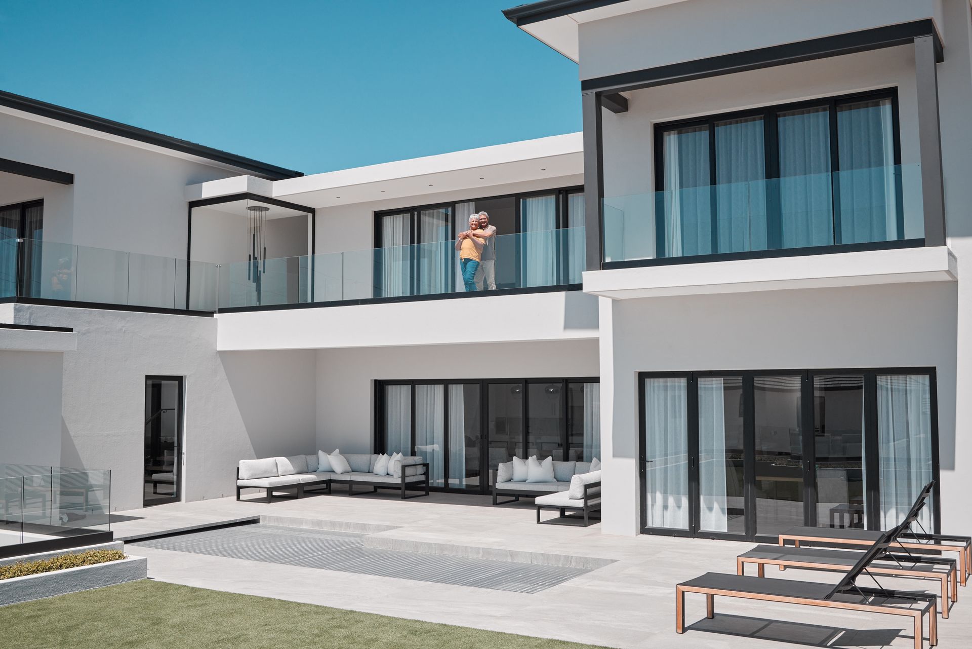 Modern white two-story house with glass doors, patio lounge chairs, and a person on the balcony