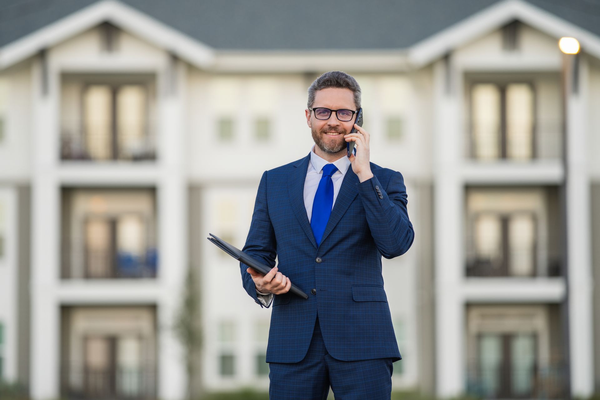 Man in a navy suit speaking on a phone while holding a tablet outside a modern apartment building
