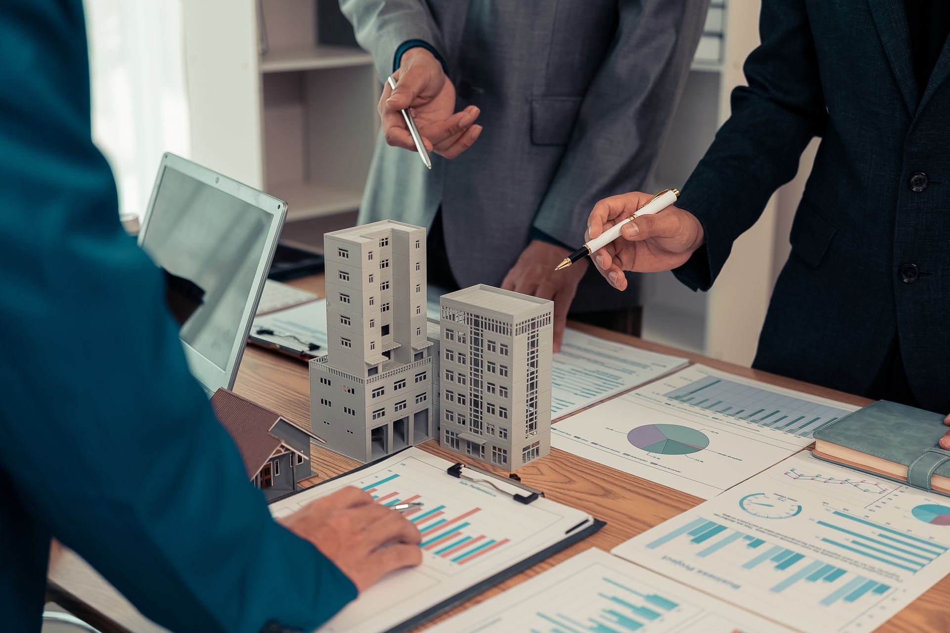 People in business attire pointing at architectural model and charts during a meeting