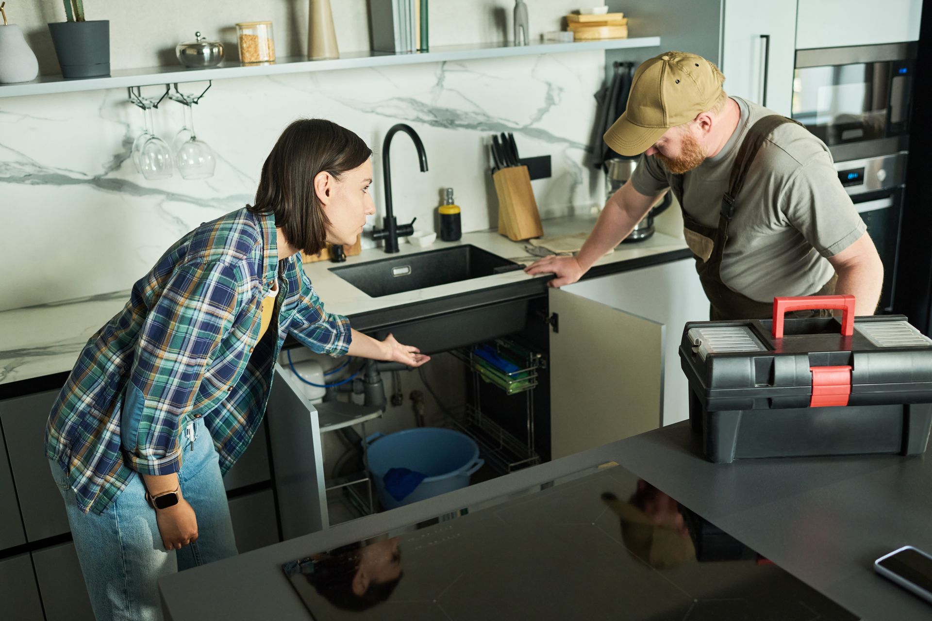 Two people inspecting a kitchen sink with tools on a black countertop