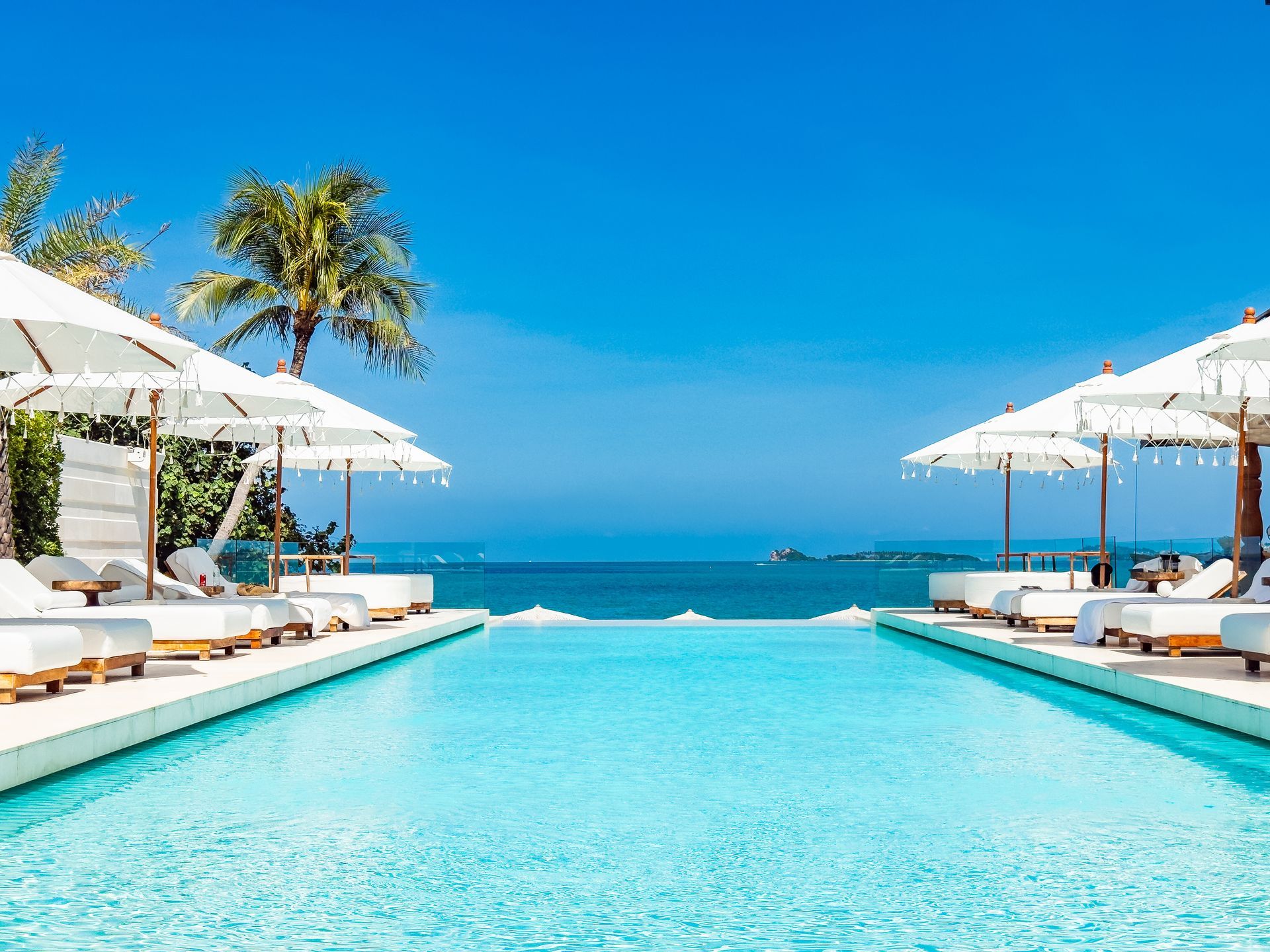 A bright, turquoise infinity pool surrounded by white lounge chairs and umbrellas, overlooking the blue ocean and sky. A bright, turquoise infinity pool surrounded by white lounge chairs and umbrellas, overlooking the blue ocean and sky.