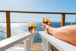 A person sits on white chairs on a deck, holding a glass of iced amber liquid while another glass sits on the chair arm. A person sits on white chairs on a deck, holding a glass of iced amber liquid while another glass sits on the chair arm.