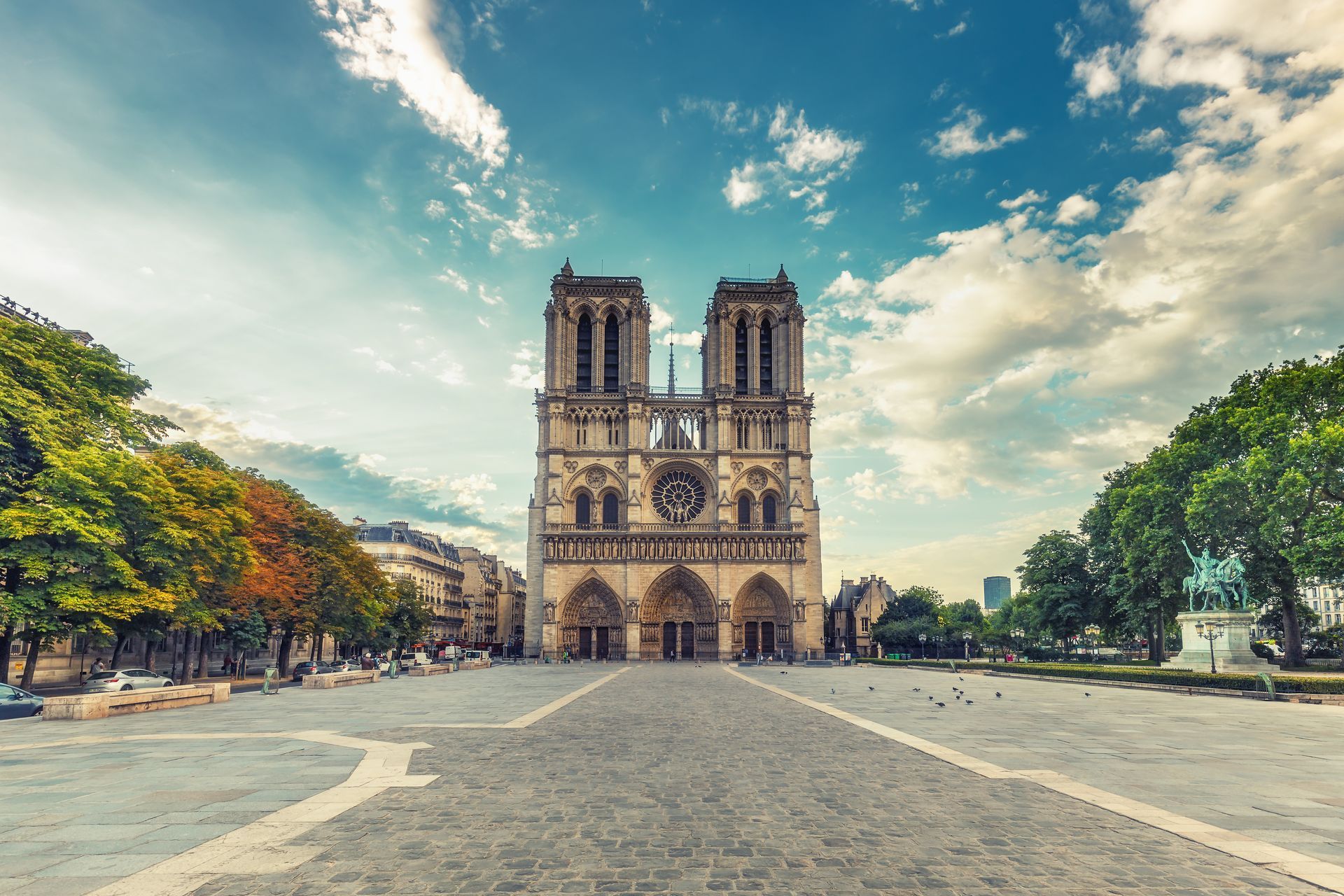 Front view of Notre-Dame Cathedral in Paris under a blue sky with scattered clouds, surrounded by trees and a stone plaza. Front view of Notre-Dame Cathedral in Paris under a blue sky with scattered clouds, surrounded by trees and a stone plaza.