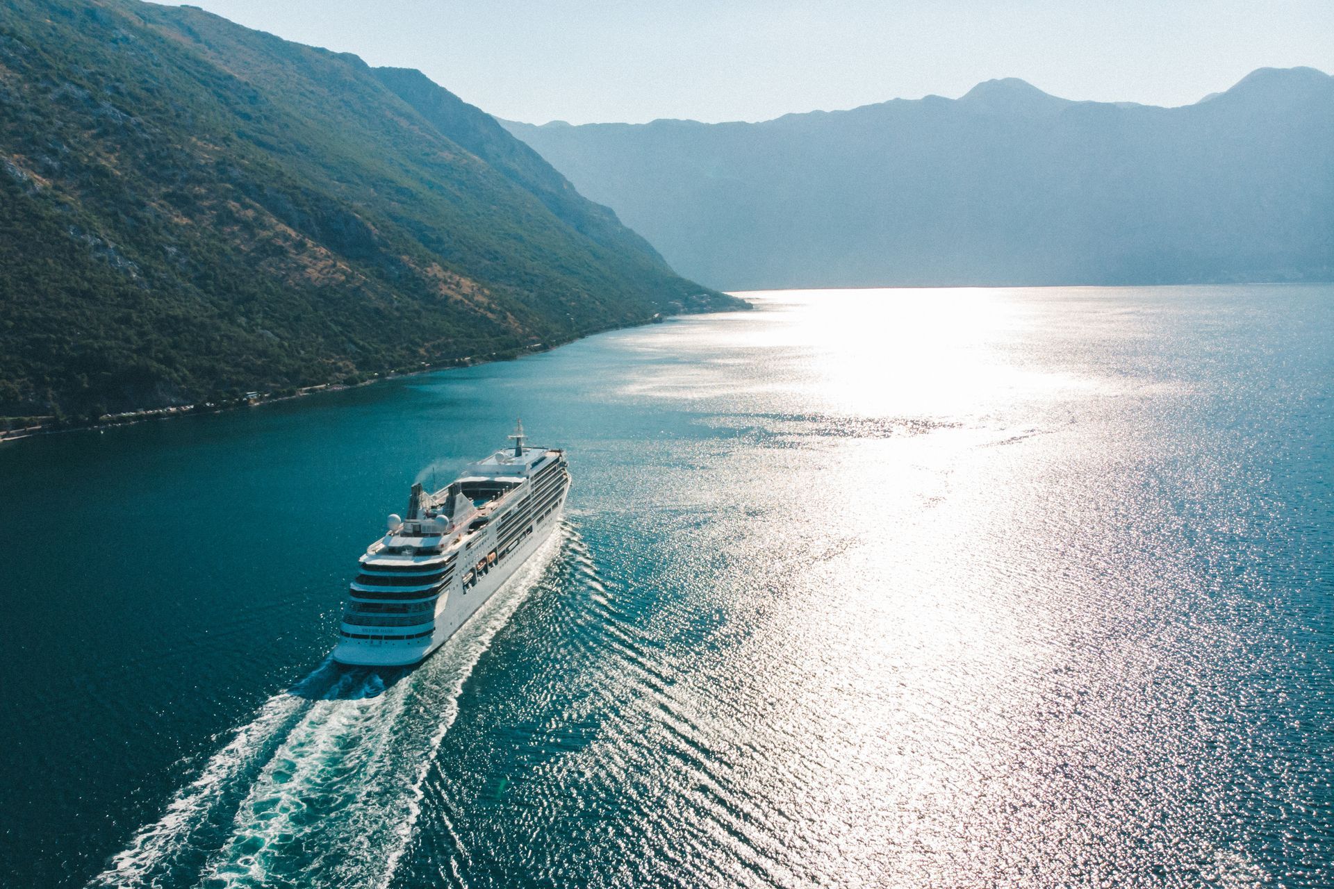 A large white cruise ship sails through a calm, sun-dappled bay surrounded by steep, grassy mountains. A large white cruise ship sails through a calm, sun-dappled bay surrounded by steep, grassy mountains.