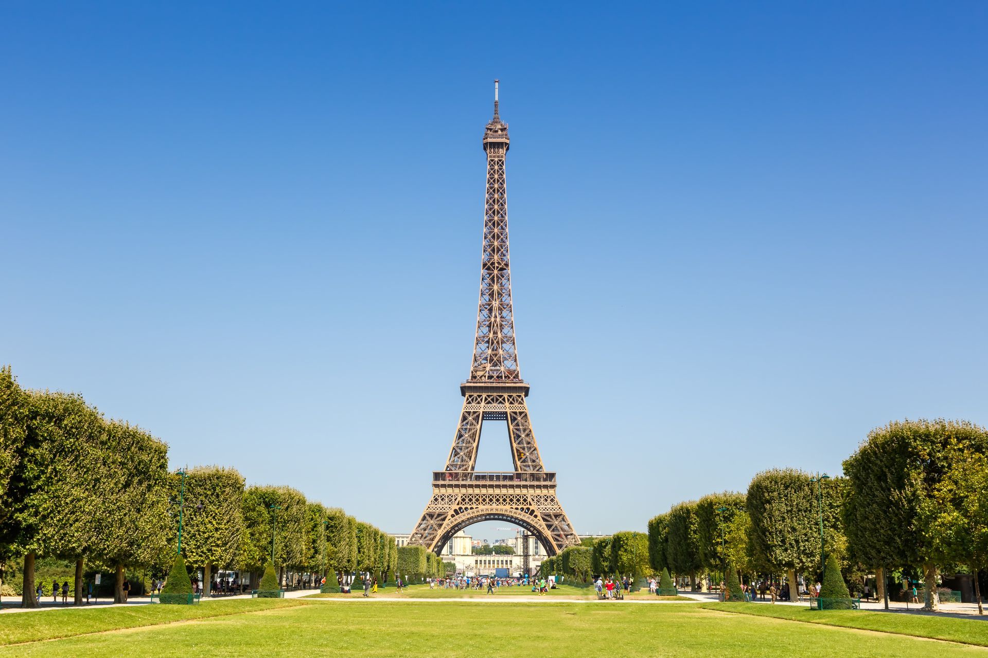 The Eiffel Tower stands in the distance, framed by manicured green trees and a lawn under a clear blue sky in Paris. The Eiffel Tower stands in the distance, framed by manicured green trees and a lawn under a clear blue sky in Paris.