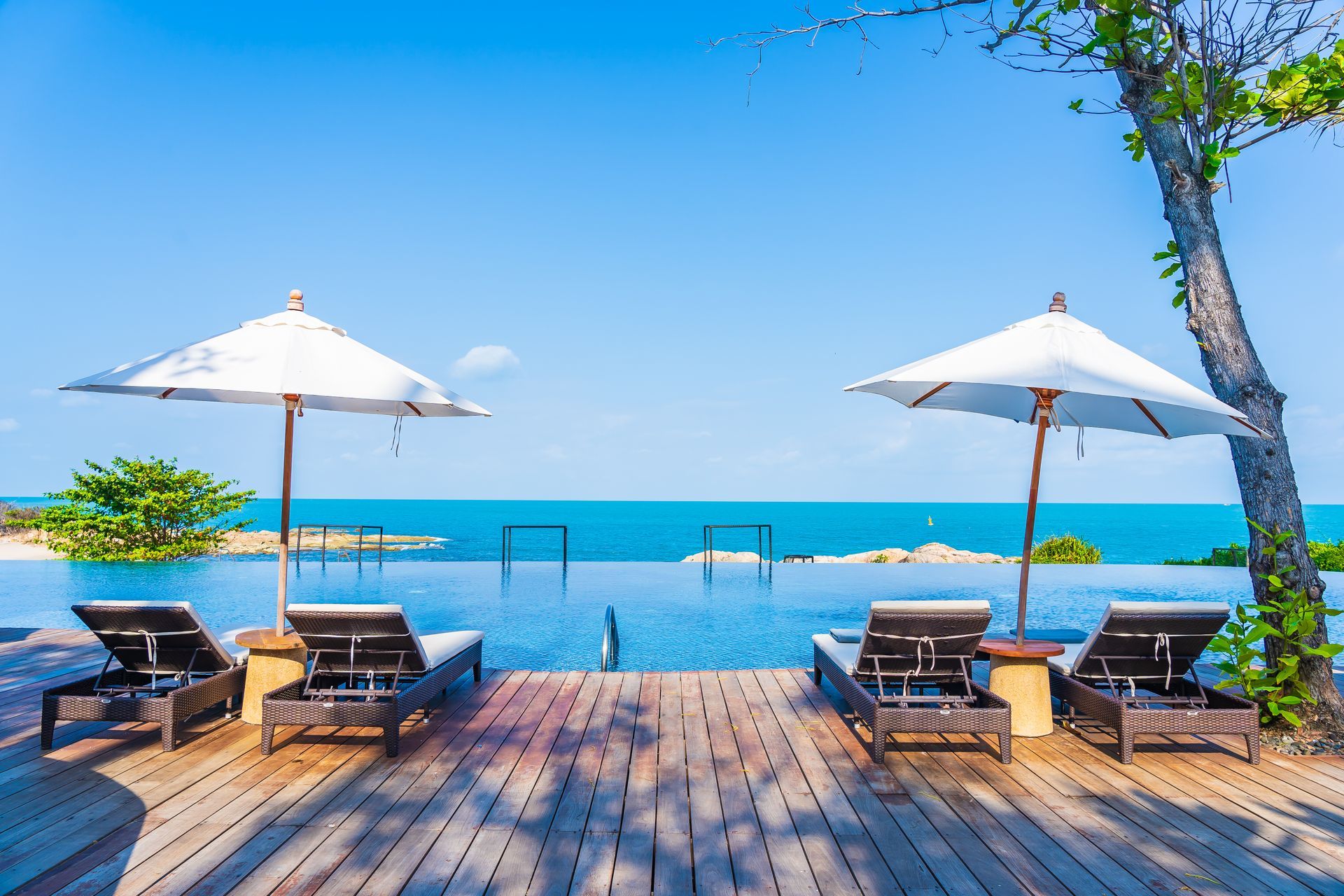 Four sun loungers with two umbrellas on a wooden deck overlooking a blue infinity pool and the sea under a clear sky. Four sun loungers with two umbrellas on a wooden deck overlooking a blue infinity pool and the sea under a clear sky.