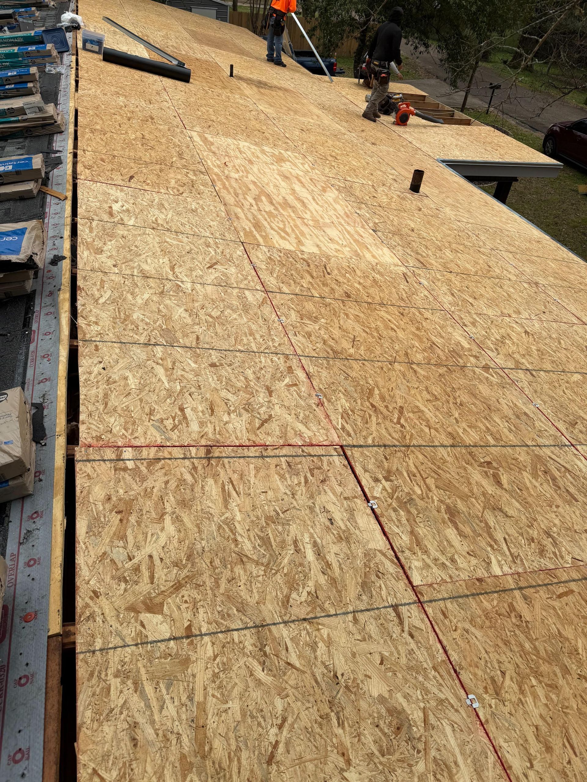 Damaged roof with torn black shingles around a white chimney on plywood decking
