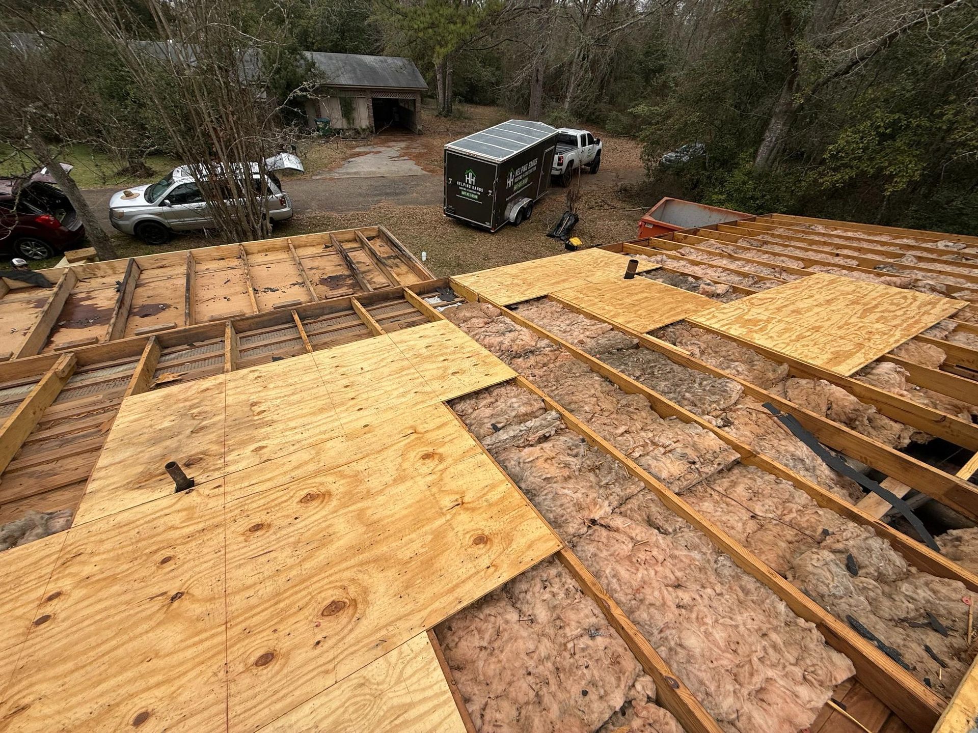 Roof decking under construction with plywood panels, workers, and vehicles on a wooded hillside.