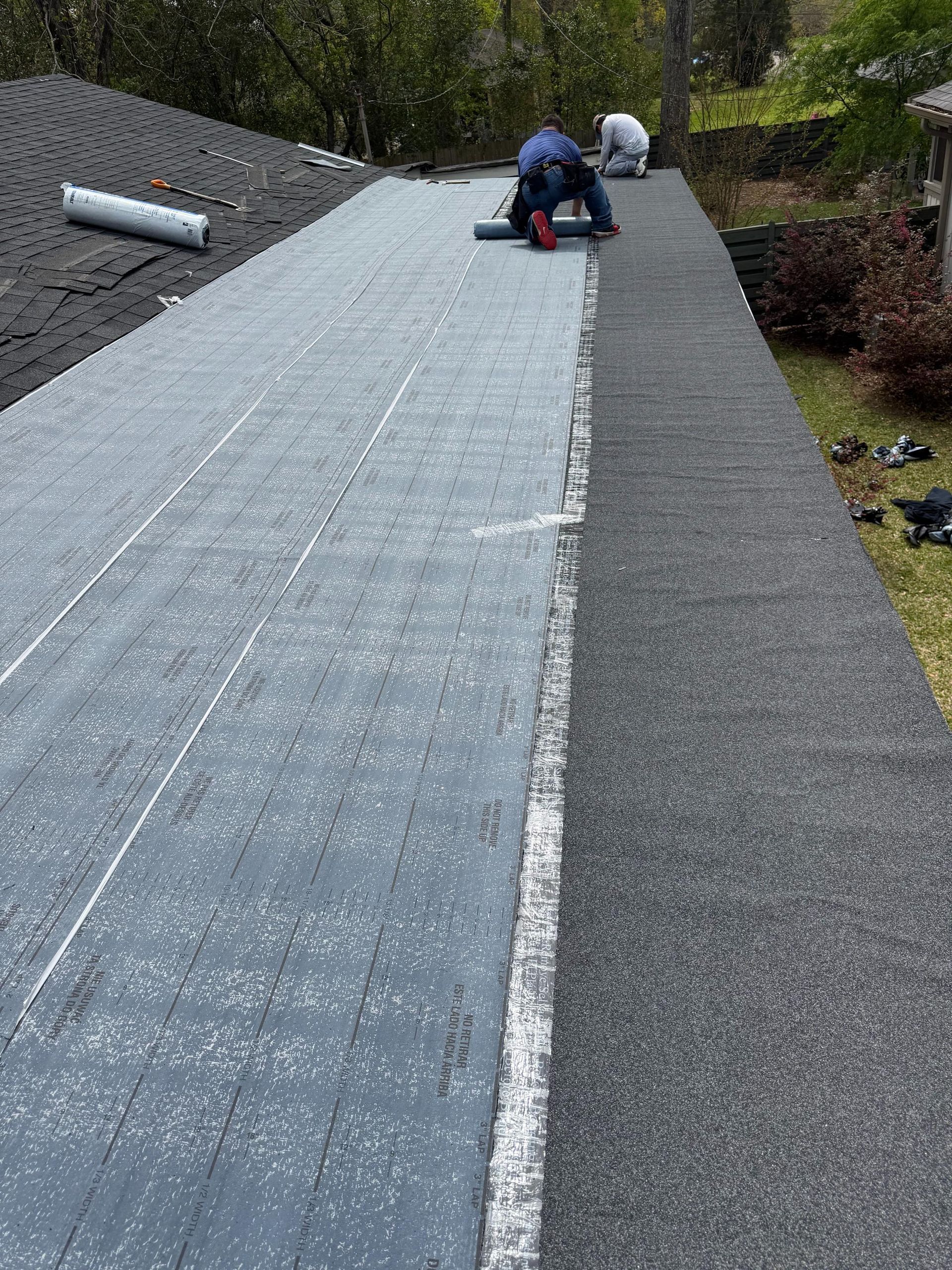 Worker installing roofing underlayment on a steep shingled roof, with rolled materials nearby.
