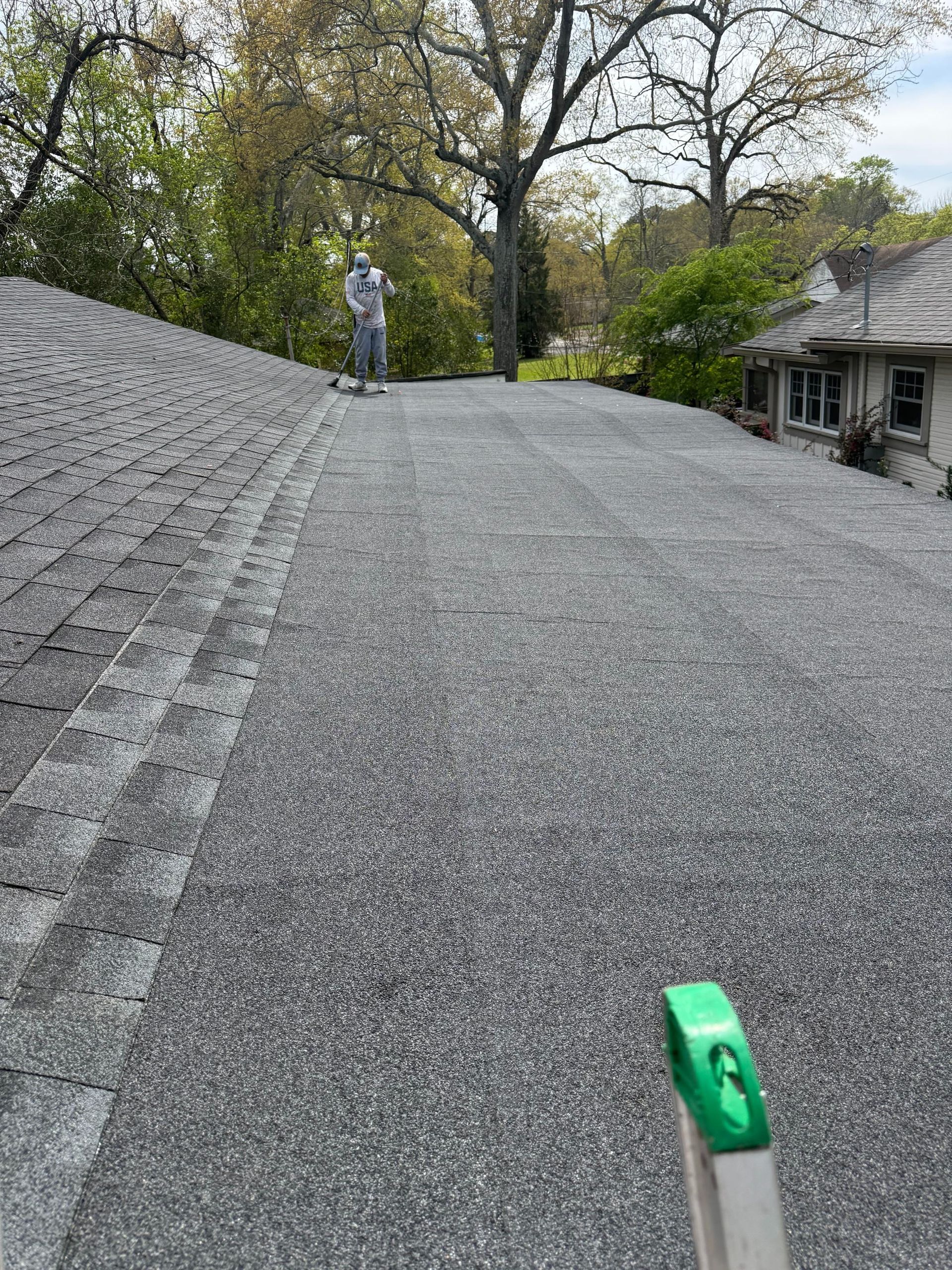Roof shingle installation with a worker standing on a sloped roof under trees.