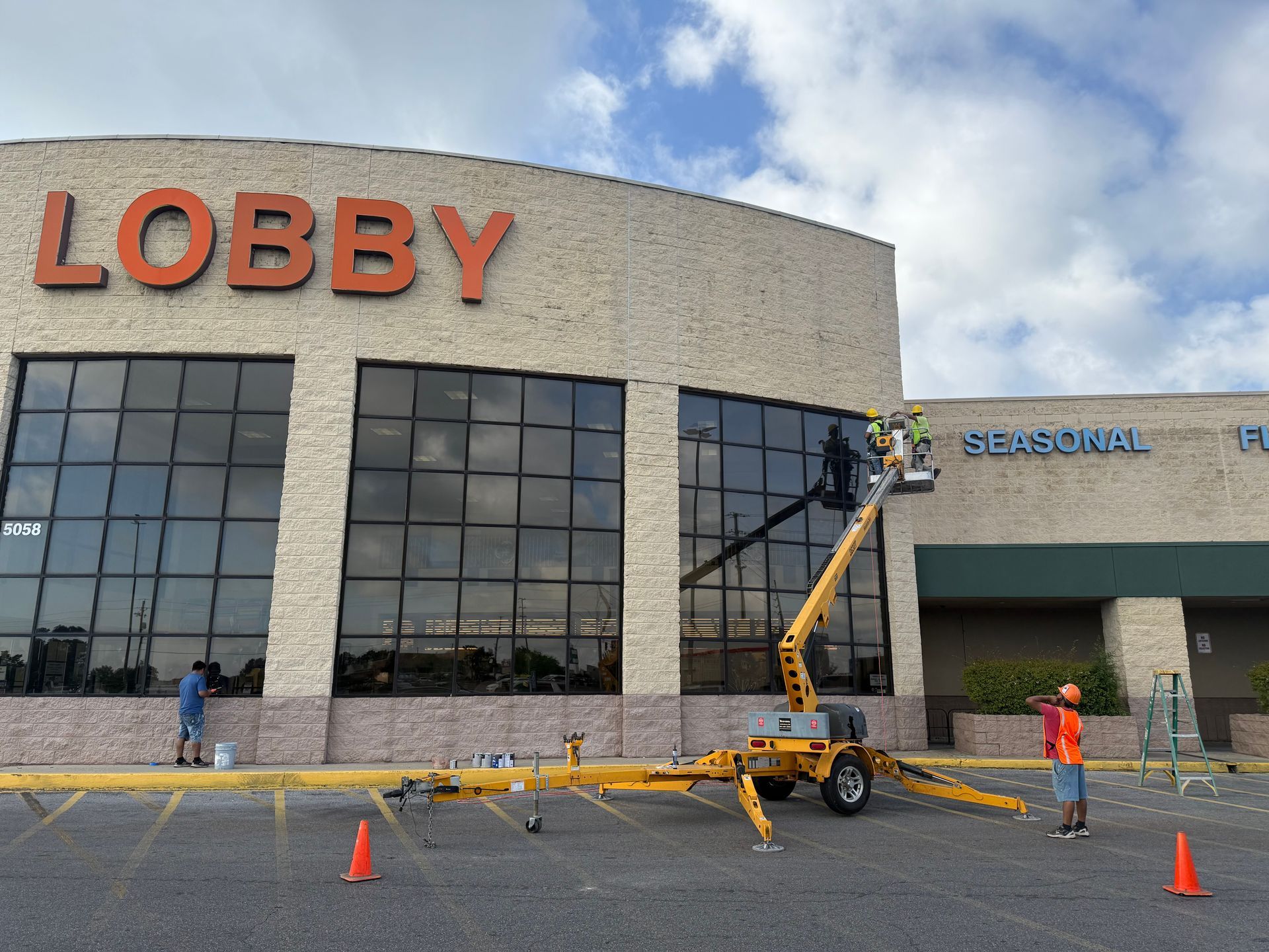 Construction workers and lift near a building labeled LOBBY, with cones and a gray facade