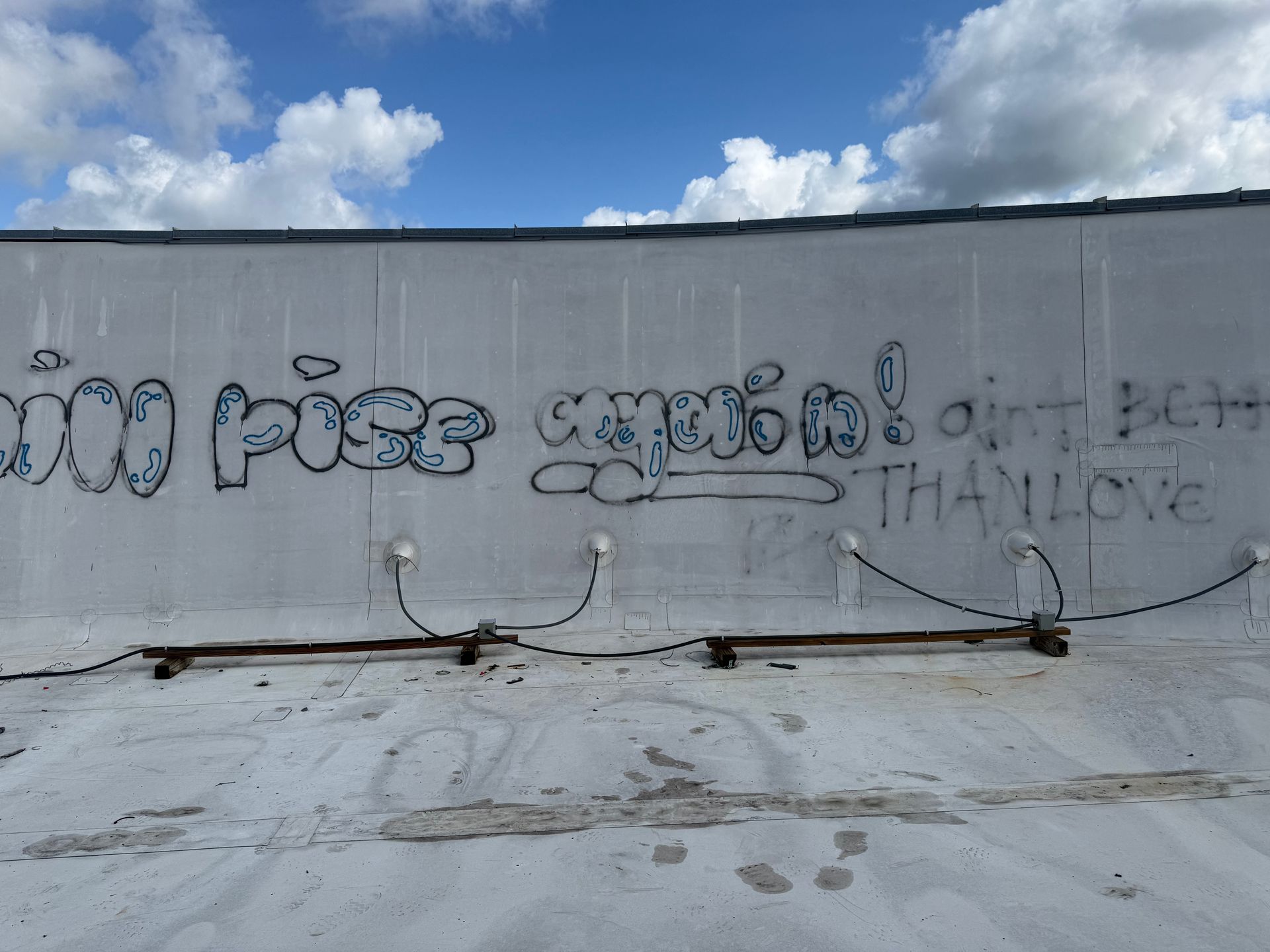 Graffiti-covered concrete wall under a blue sky with clouds, cables and pipes running along the bottom