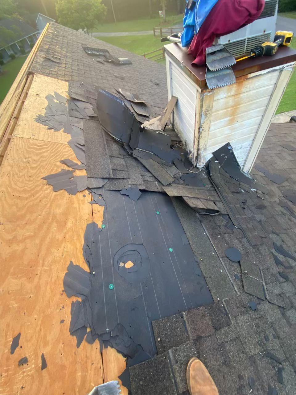 Damaged roof with torn black shingles around a white chimney on plywood decking