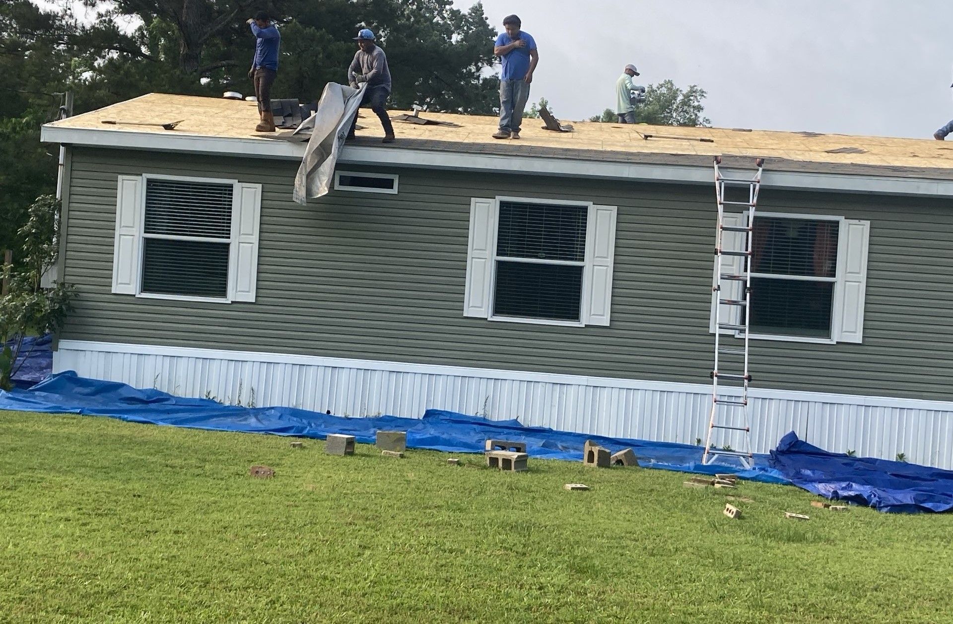 Workers installing shingles on a gray mobile home roof with blue tarps below