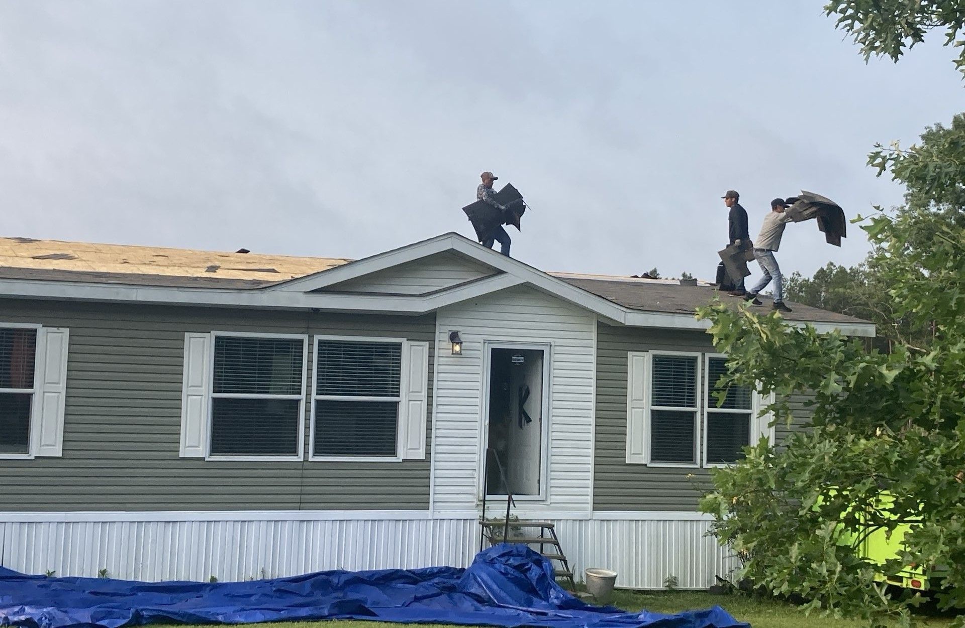 Workers repairing a mobile home roof amid storm damage, with blue tarp covering the ground