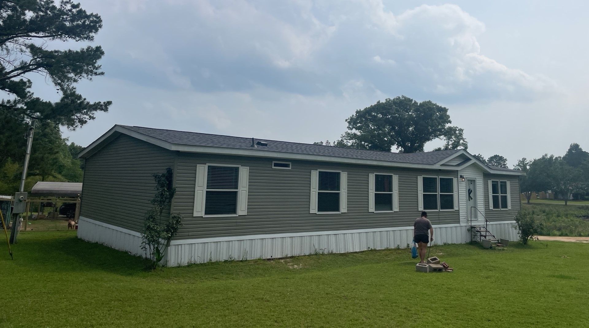 Long gray house with white trim and a white skirting, standing on a grassy lawn under a cloudy sky