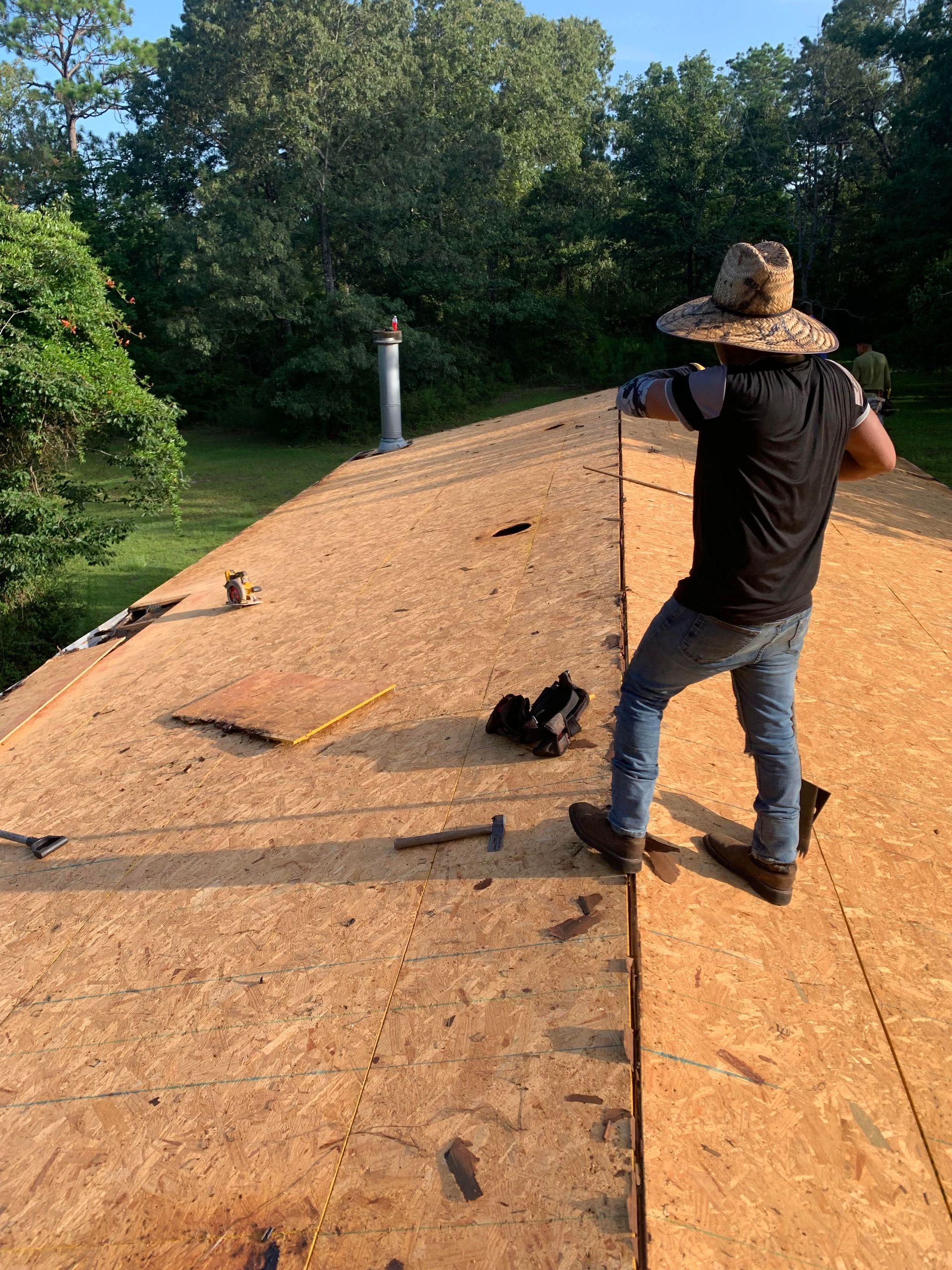 Person on a roof installing materials, with trees and blue sky in the background.