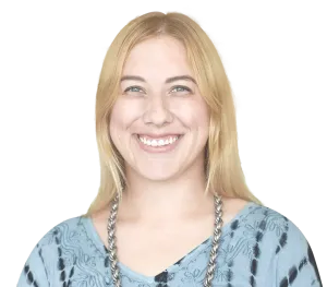 A smiling person with shoulder-length blonde hair, wearing a blue tie-dye shirt and a silver chain necklace.