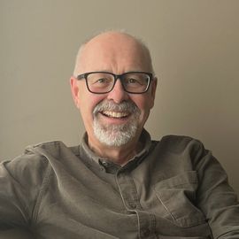 A smiling person with a goatee and glasses wearing a textured gray button-up shirt over a black shirt.