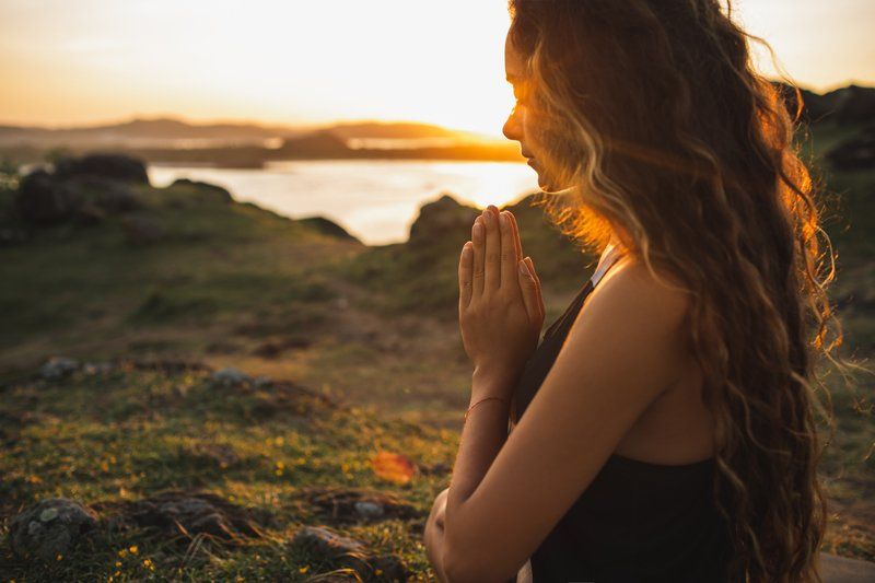 A woman is praying in front of a body of water at sunset.