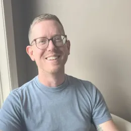 A smiling person wearing glasses and a blue t-shirt, posed indoors against a neutral, plain wall.