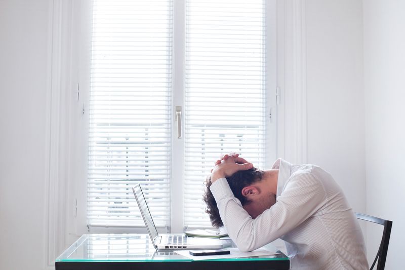 A man is sitting at a desk with his head in his hands in front of a laptop computer.