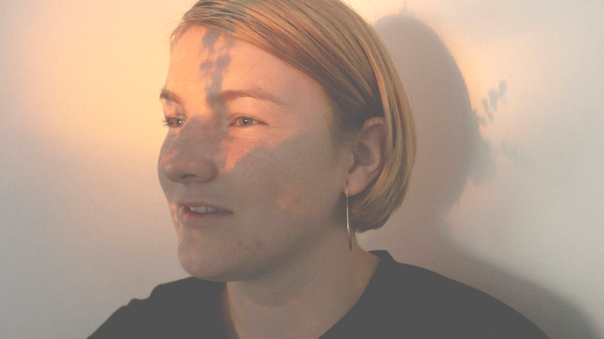 A person with short hair sits in a corner, illuminated by warm, golden sunlight casting a soft shadow on the white wall.