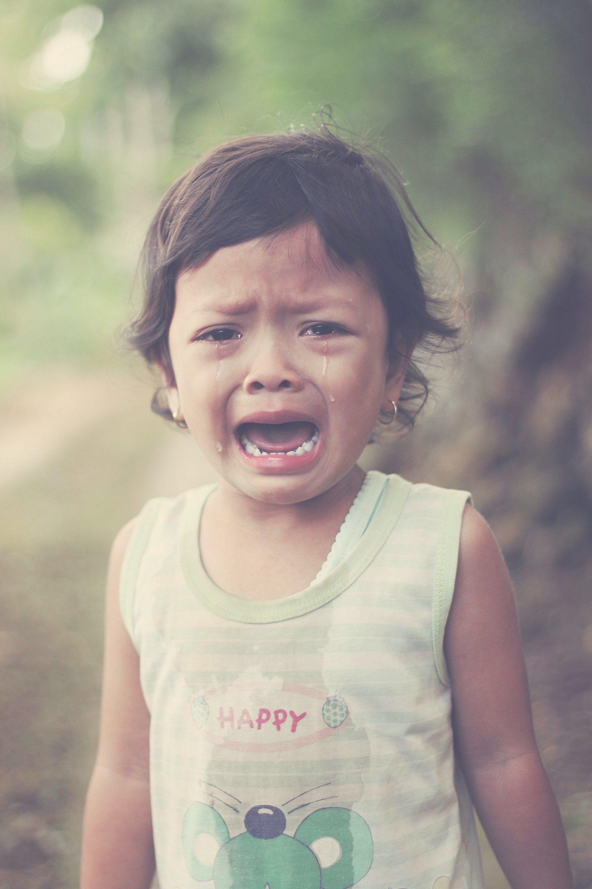 Crying toddler, wearing a green tank top, with an open mouth and tears. Outdoors, blurred background.