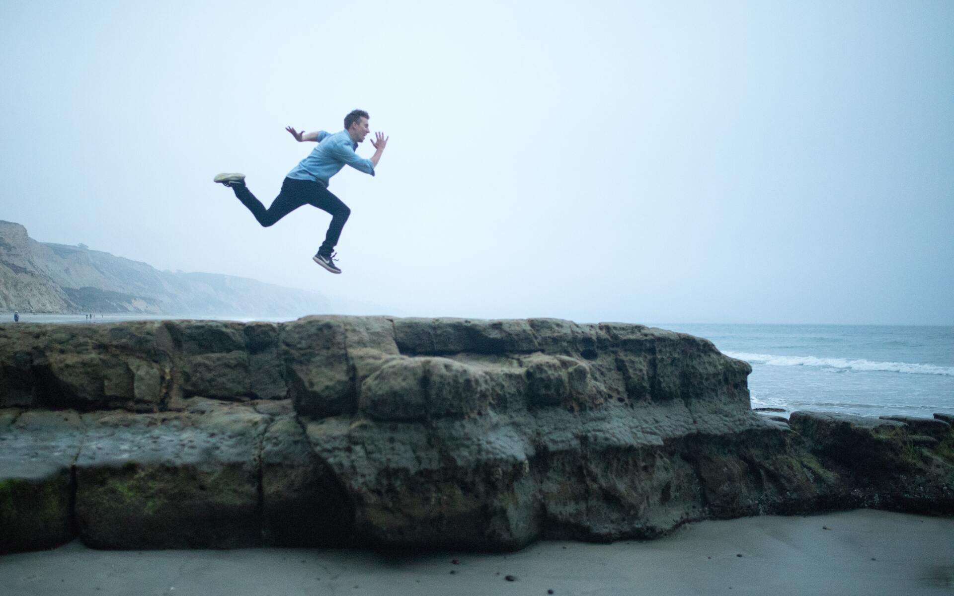A man is jumping in the air over a rocky cliff near the ocean.