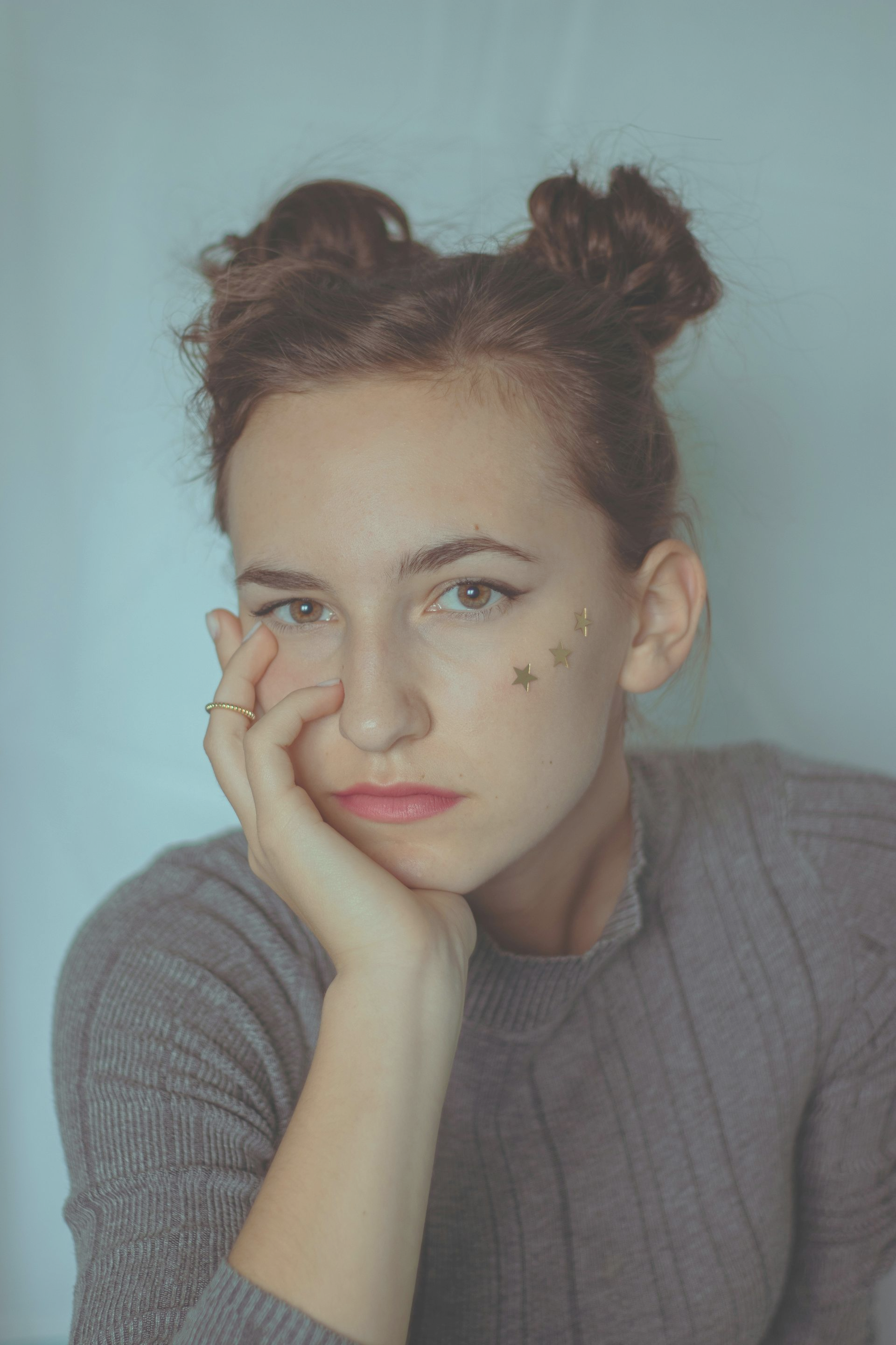 Young woman with brown hair in two buns and gray turtleneck shirt sits slumped with hand under chin.
