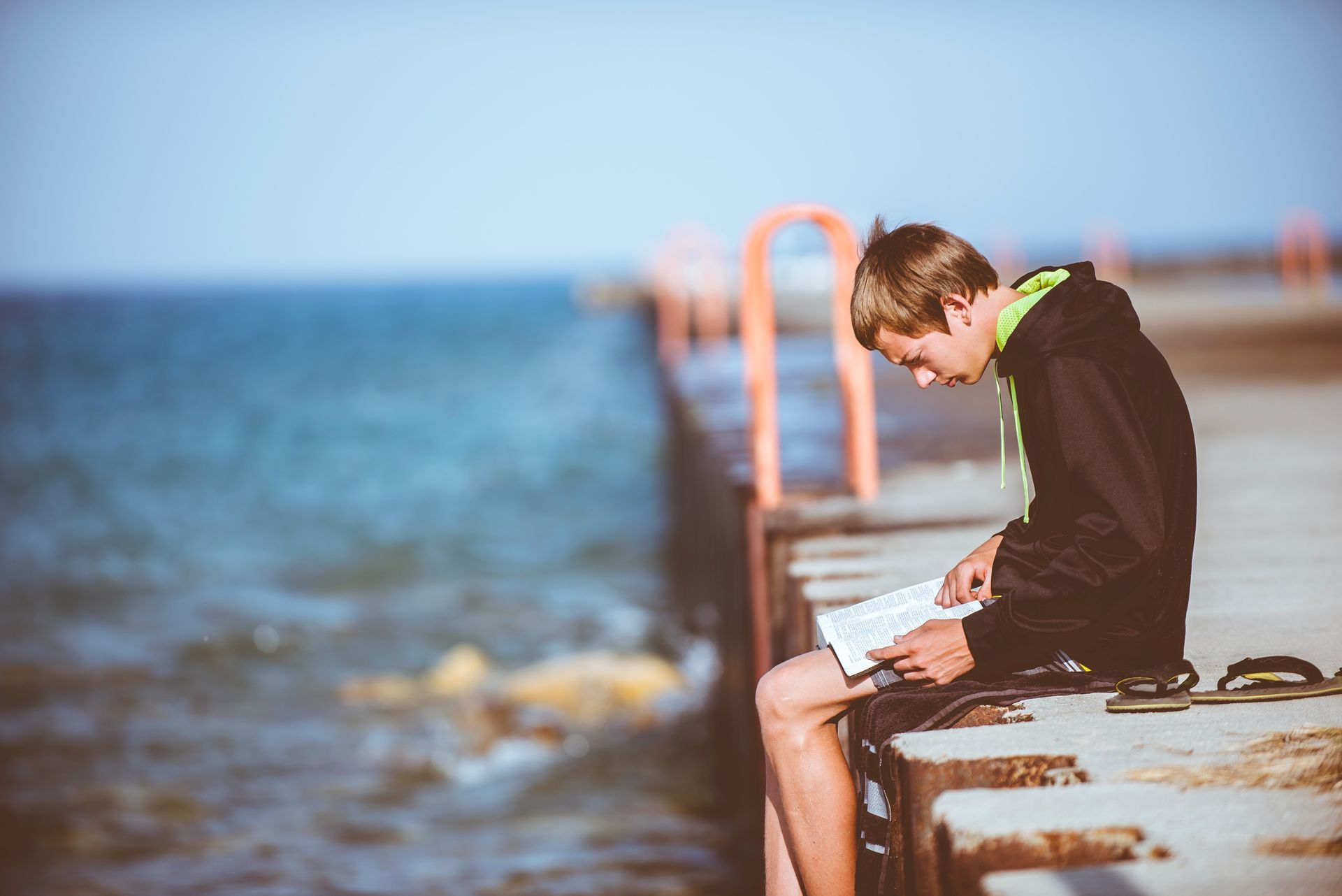 A young man is sitting on a pier reading a book.