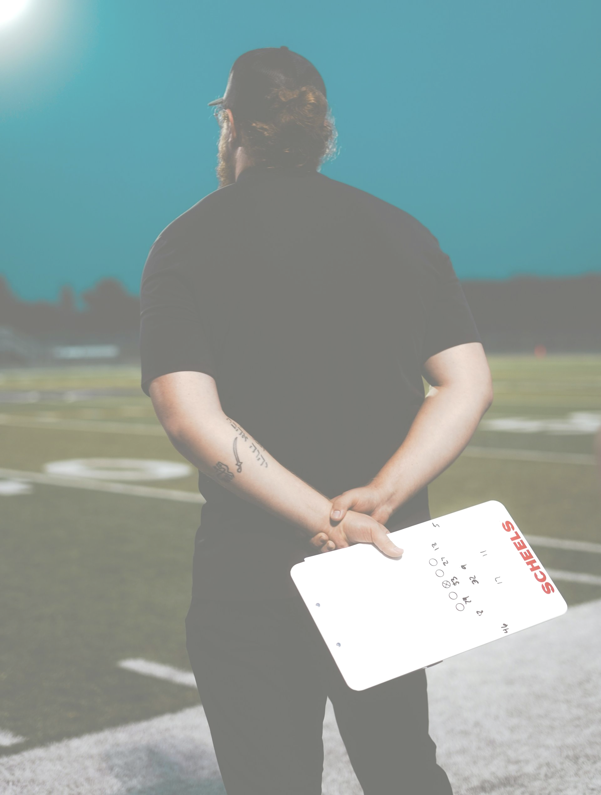 A person with long hair tied back stands on a football field, back to the camera, holding a blank white clipboard.
