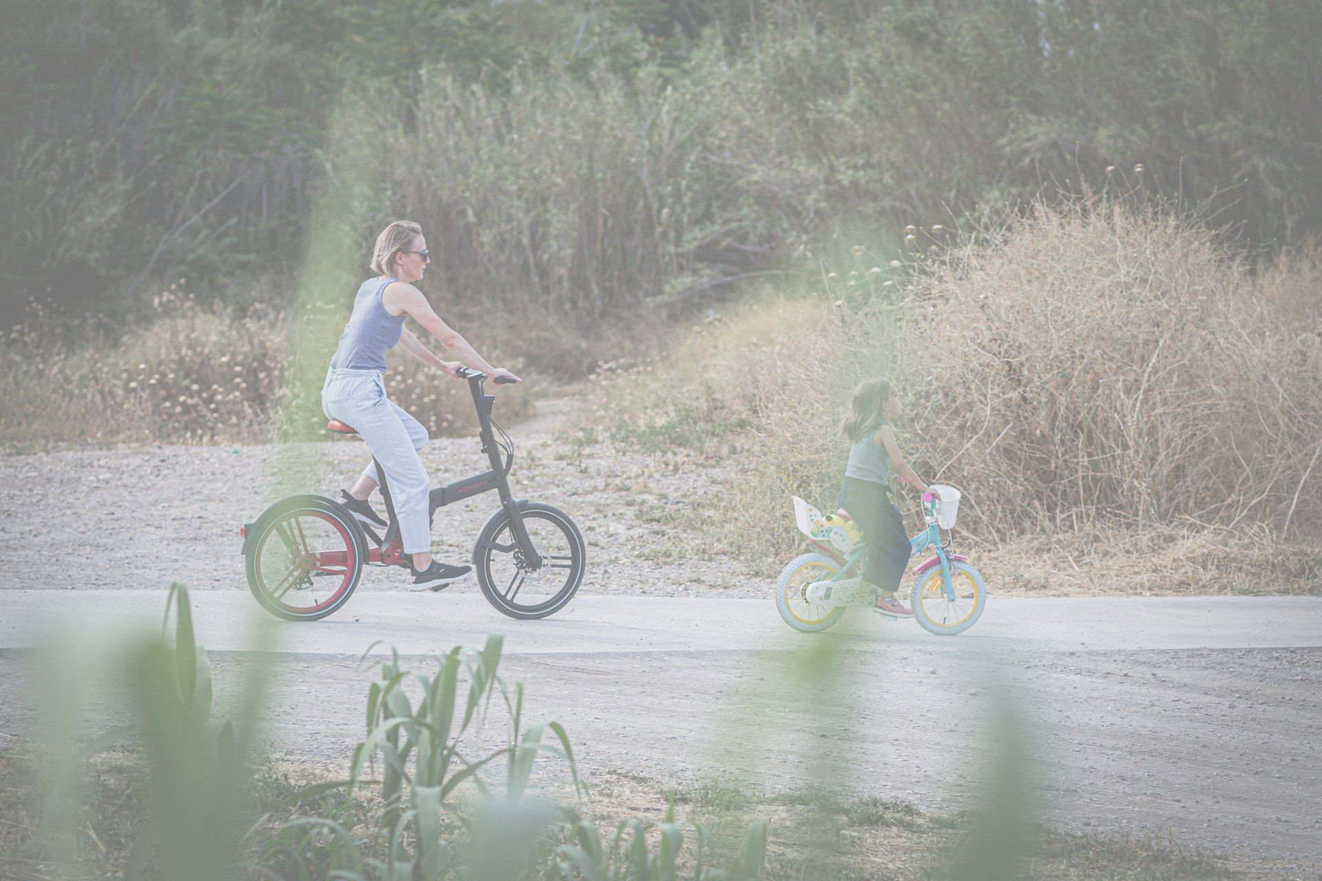 Woman on black bicycle, child on scooter, both on path by grassy roadside.