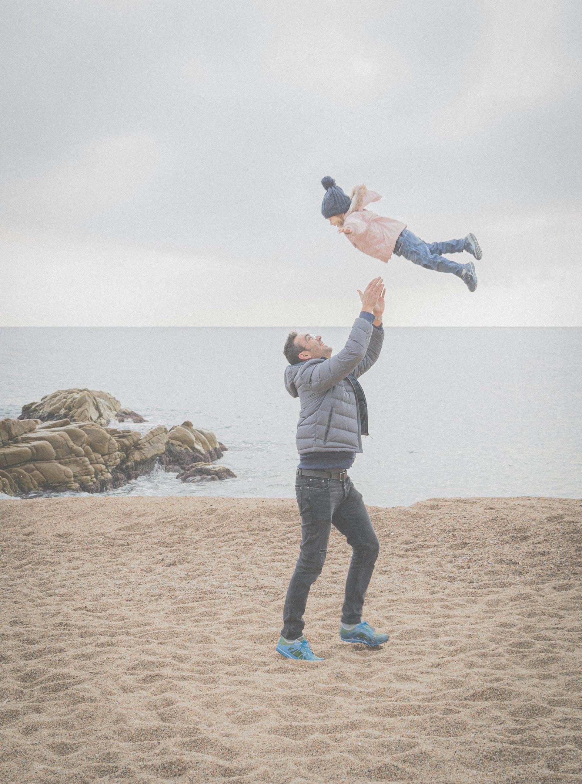 Man throws a child in the air on a sandy beach. Cloudy sky, ocean in the background.