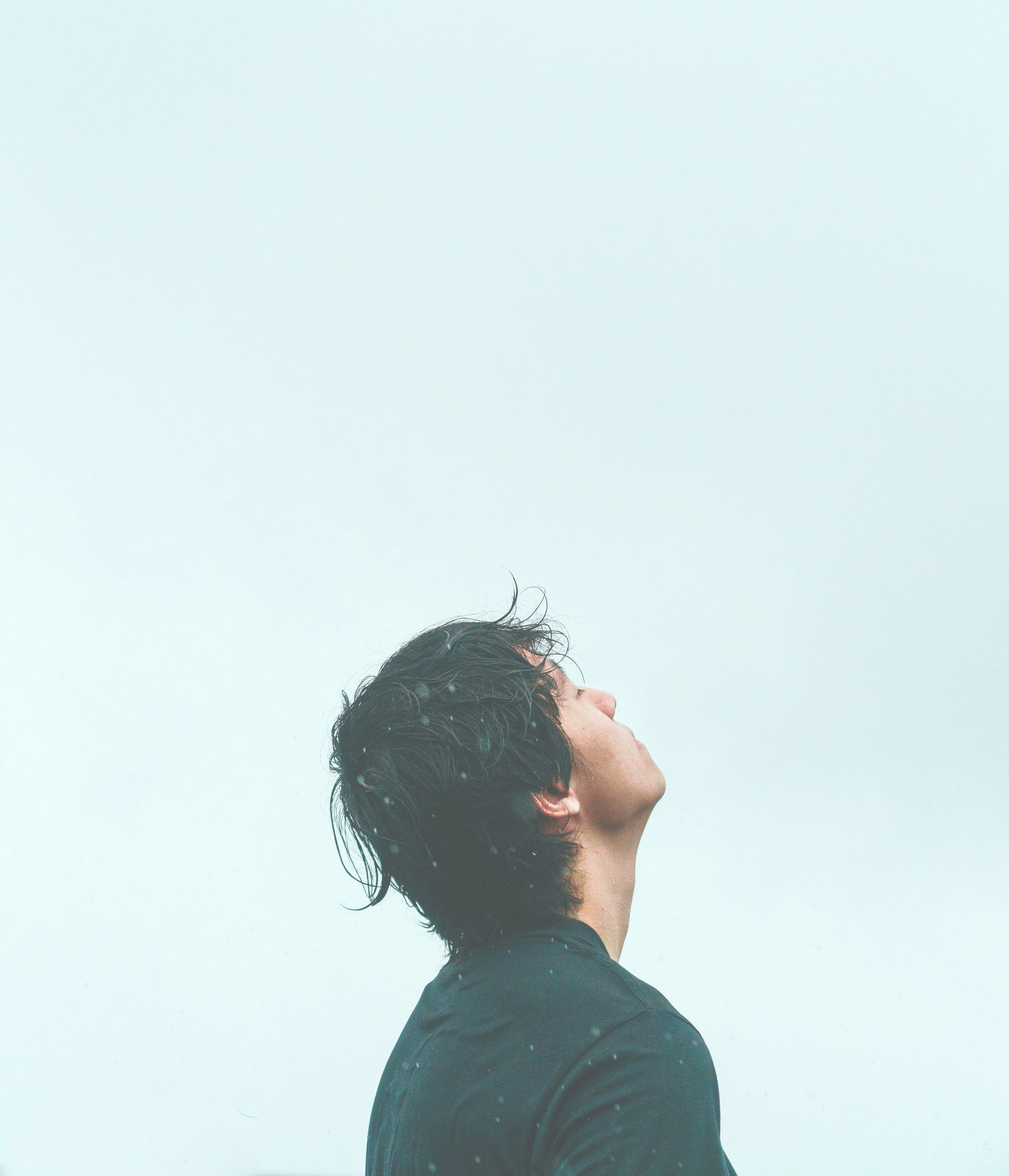 Man looking up at the sky. He has dark, curly hair and wears a black shirt against a cloudy, light backdrop.
