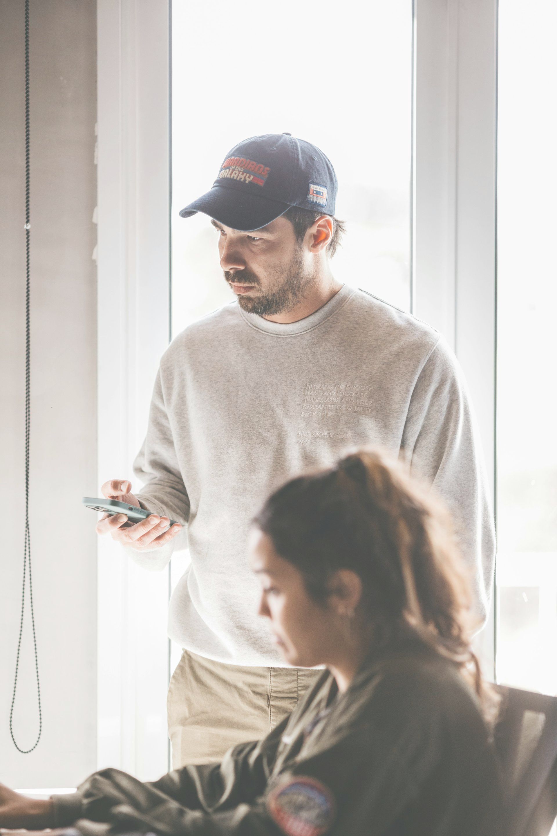 Man in a baseball cap looks at a phone, while a woman sits at a desk in front of a window.