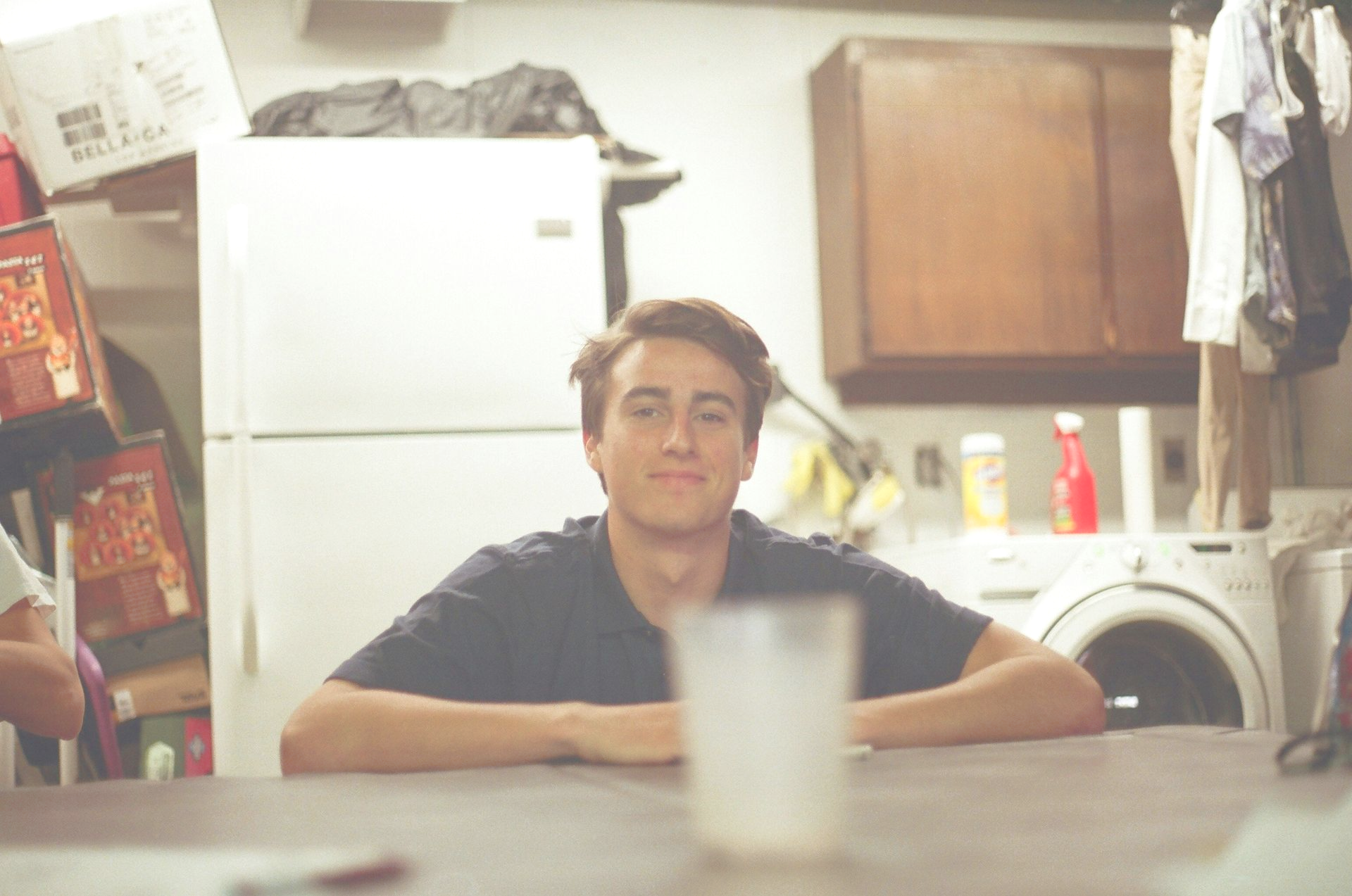 Young man sitting at a table smiling, with a washing machine and storage in the background.