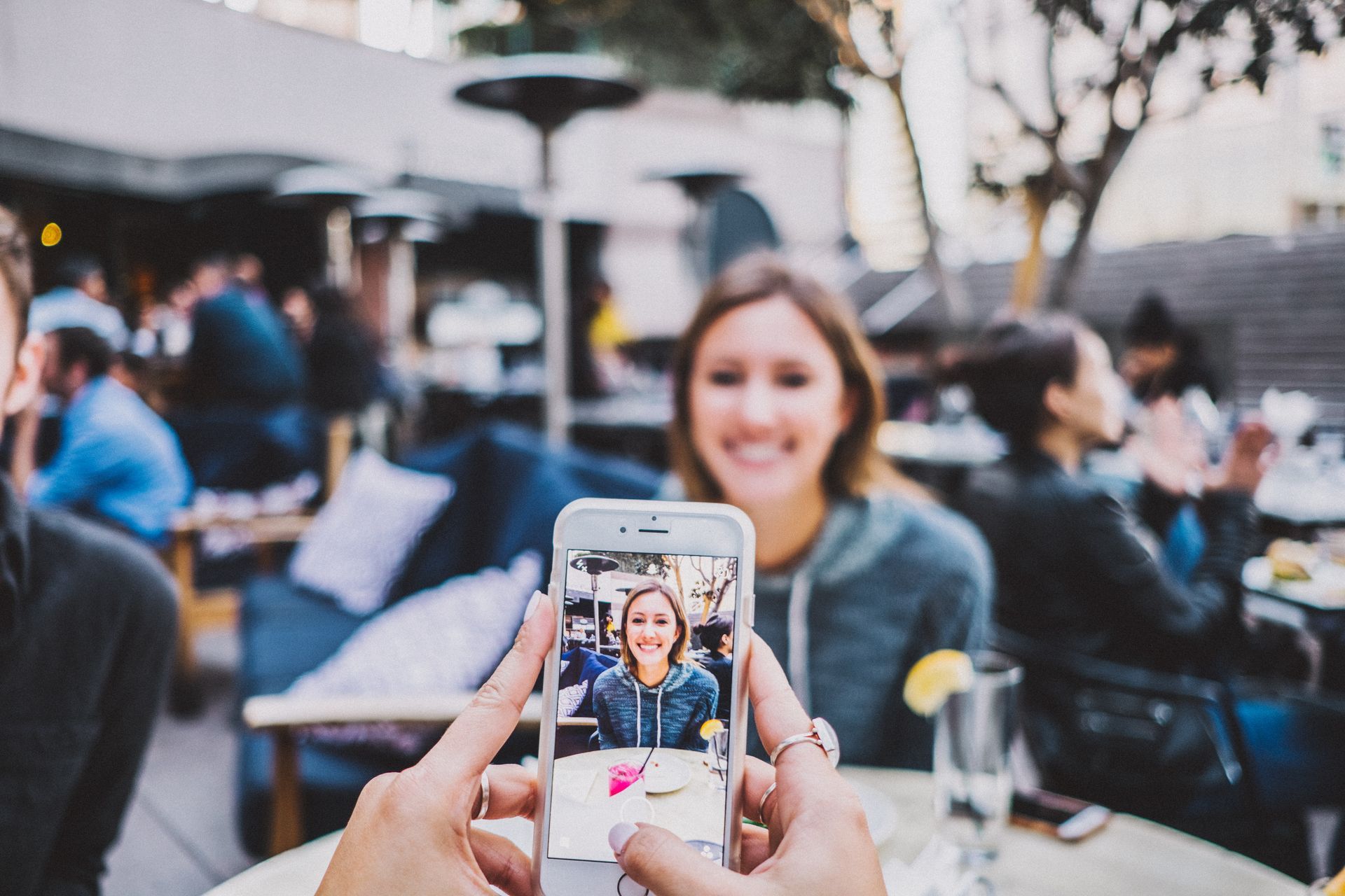 A person is taking a picture of a woman sitting at a table with a cell phone.