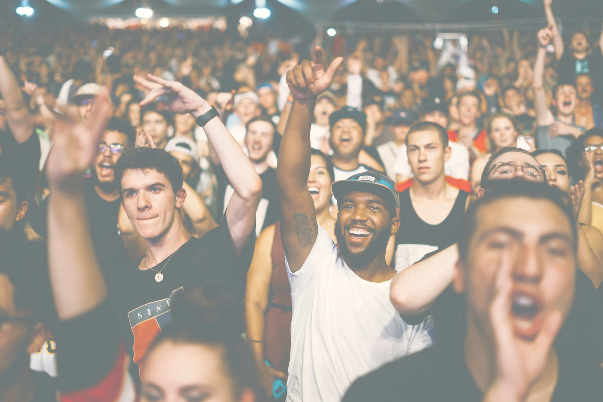 Excited concert crowd with arms raised, cheering and smiling, under bright lights.