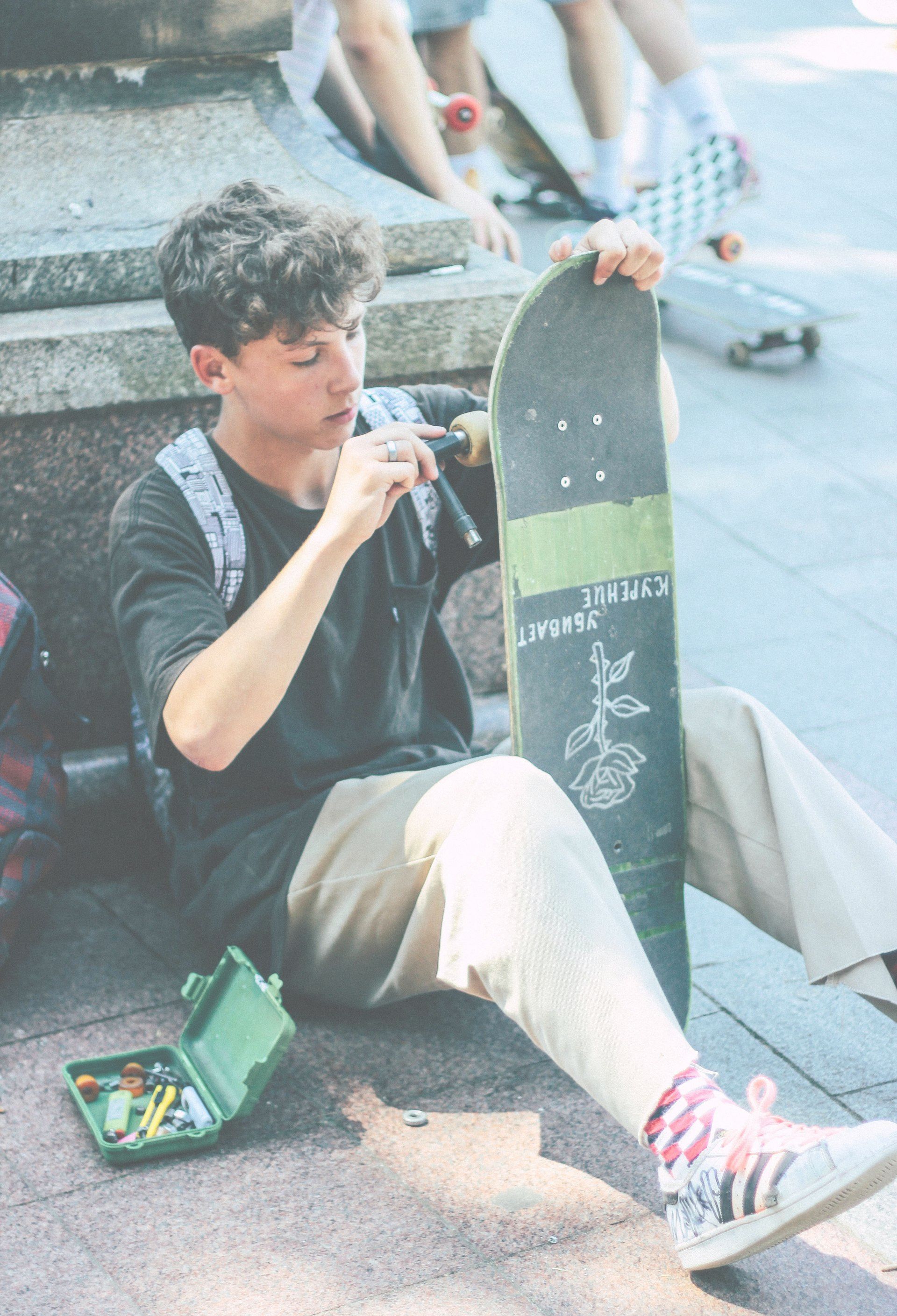 Teenager sits on ground, fixing skateboard, with open tool case. City setting.